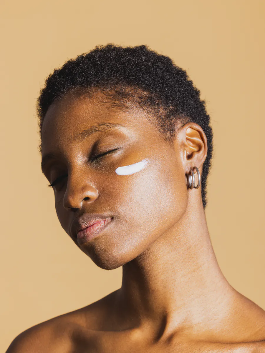 A model with glowing skin and a stroke of cream on one cheek, eyes closed, on a warm tan studio field.