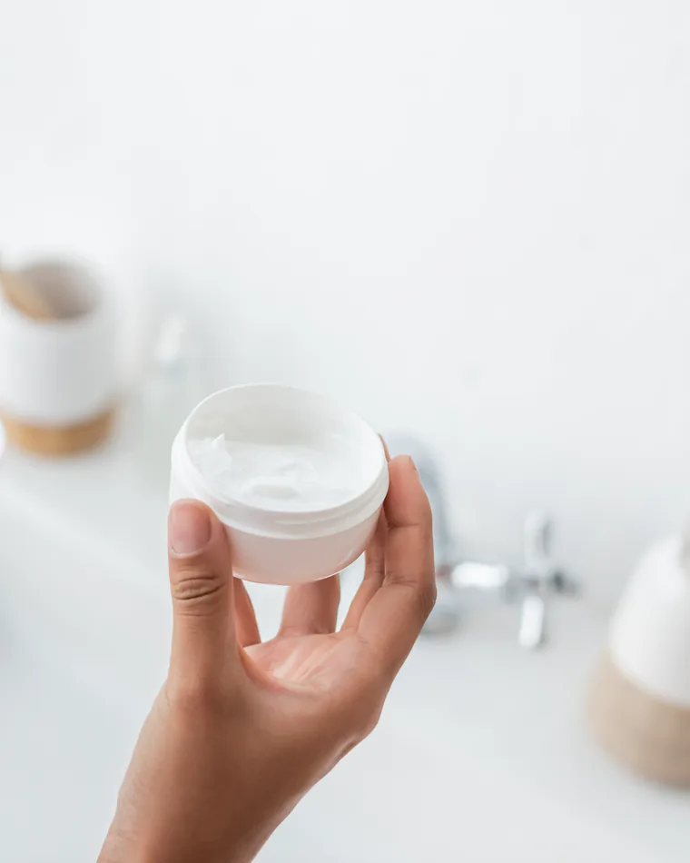Hands holding an open jar of skin cream over a bright, clean counter.