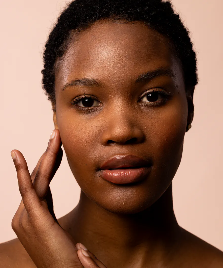 A model resting a hand against her cheek, soft glossy skin, on a warm blush studio field.